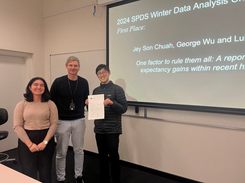 Dr Shila Ghazanfar and Dr Alistair Senior stand next to Jeyson who is holding a certificate. They are standing in front of a screen that has the title of Jeyson's team entry written on it.  